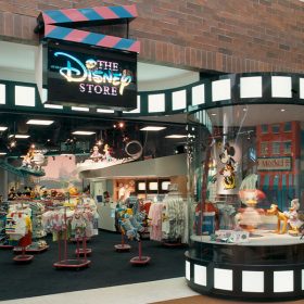 The outside of The Disney Store is shown with a black clapboard sign that features a pink and blue top clapper and has The Disney Store written on it in pink and blue alternating colors. The store's exterior is rimmed with film strips at the top and bottom. The display window features Daisy Duck and the inside of the store is filled with clothes and stuffed animals.