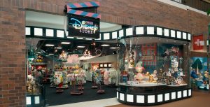 The outside of The Disney Store is shown with a black clapboard sign that features a pink and blue top clapper and has The Disney Store written on it in pink and blue alternating colors. The store's exterior is rimmed with film strips at the top and bottom. The display window features Daisy Duck and the inside of the store is filled with clothes and stuffed animals.