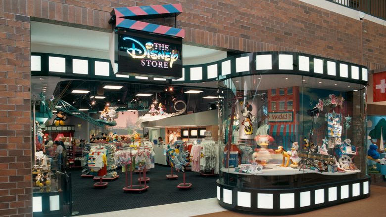 The outside of The Disney Store is shown with a black clapboard sign that features a pink and blue top clapper and has The Disney Store written on it in pink and blue alternating colors. The store's exterior is rimmed with film strips at the top and bottom. The display window features Daisy Duck and the inside of the store is filled with clothes and stuffed animals.