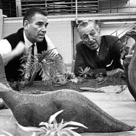 Disney Legend and Imagineer Claude Coats, on the left, consults with Walt Disney, on the right, behind a tabletop scale model of a scene with two large Brontosauruses with their offspring standing in water that was planned for the Magic Skyway attraction at the 1964–1965 New York World’s Fair. In the black-and-white image, Claude wears a white shirt, thin black tie, and sweater vest, while Walt wears a dark suit and tie. Both men are looking at the model dinosaurs; Claude is gesturing with his right hand.