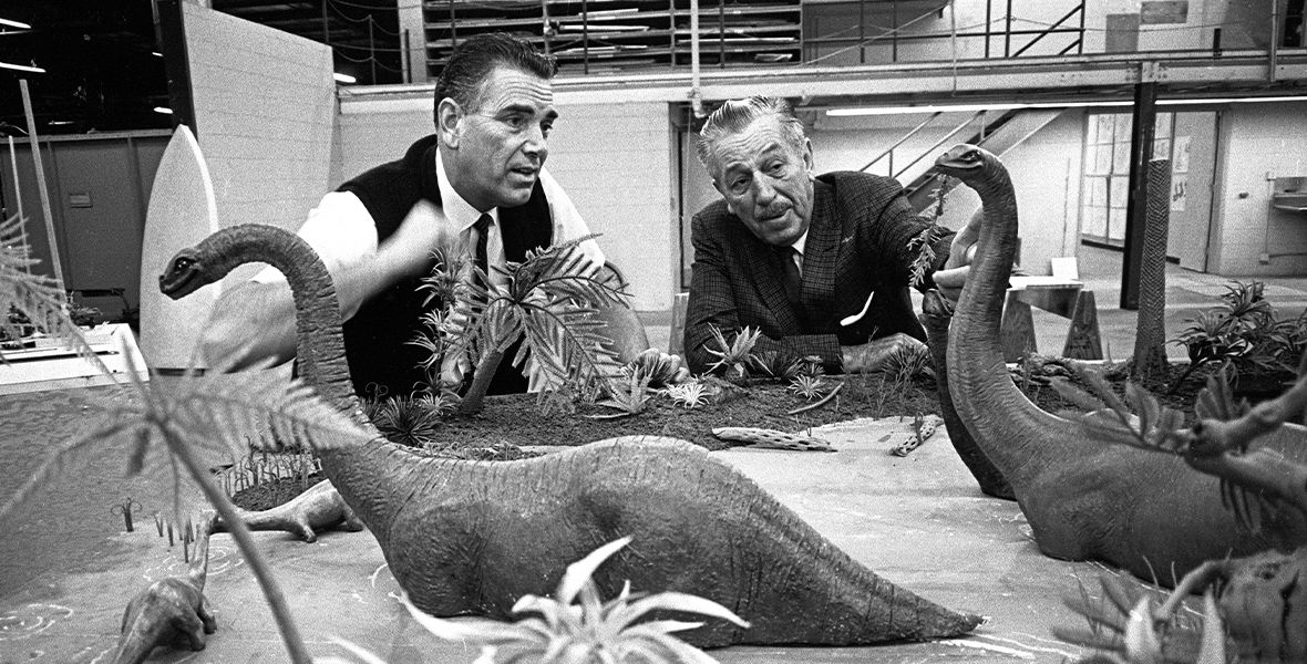 Disney Legend and Imagineer Claude Coats, on the left, consults with Walt Disney, on the right, behind a tabletop scale model of a scene with two large Brontosauruses with their offspring standing in water that was planned for the Magic Skyway attraction at the 1964–1965 New York World’s Fair. In the black-and-white image, Claude wears a white shirt, thin black tie, and sweater vest, while Walt wears a dark suit and tie. Both men are looking at the model dinosaurs; Claude is gesturing with his right hand.