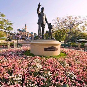 photo of statue of Walt Disney holding hands with Mickey Mouse at Disneyland, placard reading Partners, surrounded by large circular flower bed with Seeping Beauty Castle in background