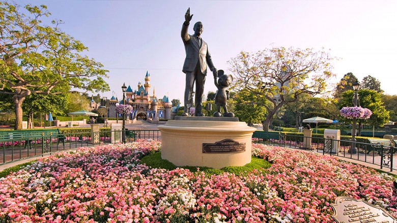 photo of statue of Walt Disney holding hands with Mickey Mouse at Disneyland, placard reading Partners, surrounded by large circular flower bed with Seeping Beauty Castle in background