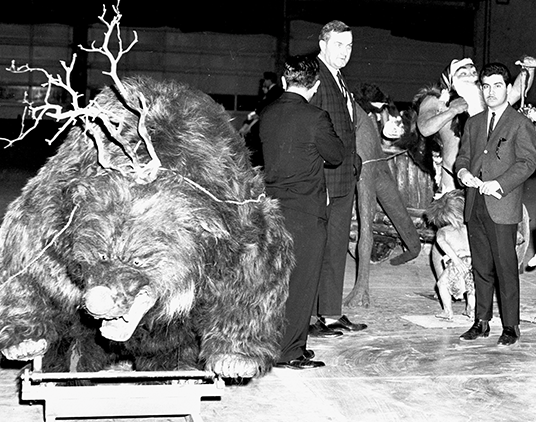 photo of huge stuffed bear-like beast with snarled antlers plus a life-size replica of a cave man and some live suit-wearing men standing bewteen