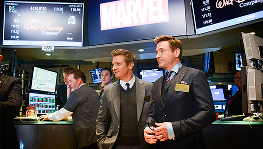 movie still of Robert Downey Jr. (Iron Man) and Jeremy Renner (Hawkeye) at New York Stock exchange wearing suits beneath stock price displays for The Walt Disney Company and Marvel