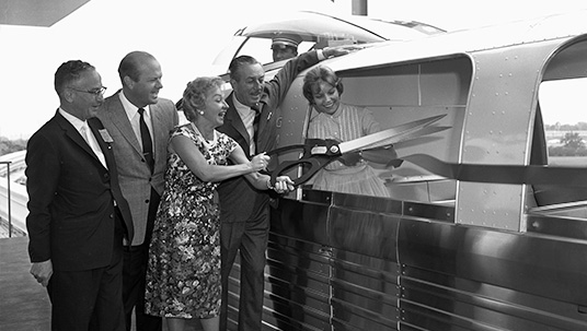 photo of Walt Disney officiating the dedication of the Monorail-to-Disneyland Hotel extension, accompanied by California Congressman James Utt as actress Bonita Granville wields giant scissors to cut the ribbon as her husband, Disneyland Hotel owner and president Jack Wrather, watches. On the Monorail is Mary Boggess of Kentucky, the first passenger to ride the newly expanded route.