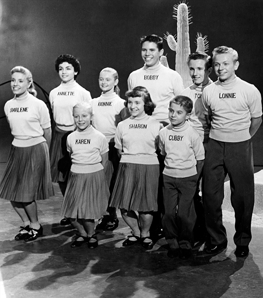 photo of nine original Mouseketeers standing in two rows bending knees in a dance step with a cactus behind them