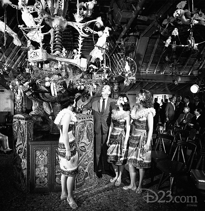 black and white photo of Walt Disney with three tropically dressed ladies inside the enchanted tiki room looking up at the central hanging display