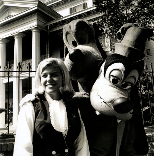 black and white photo of young Shari Bescos Koch posing with Goofy in front of Disneyland's Haunted Mansion