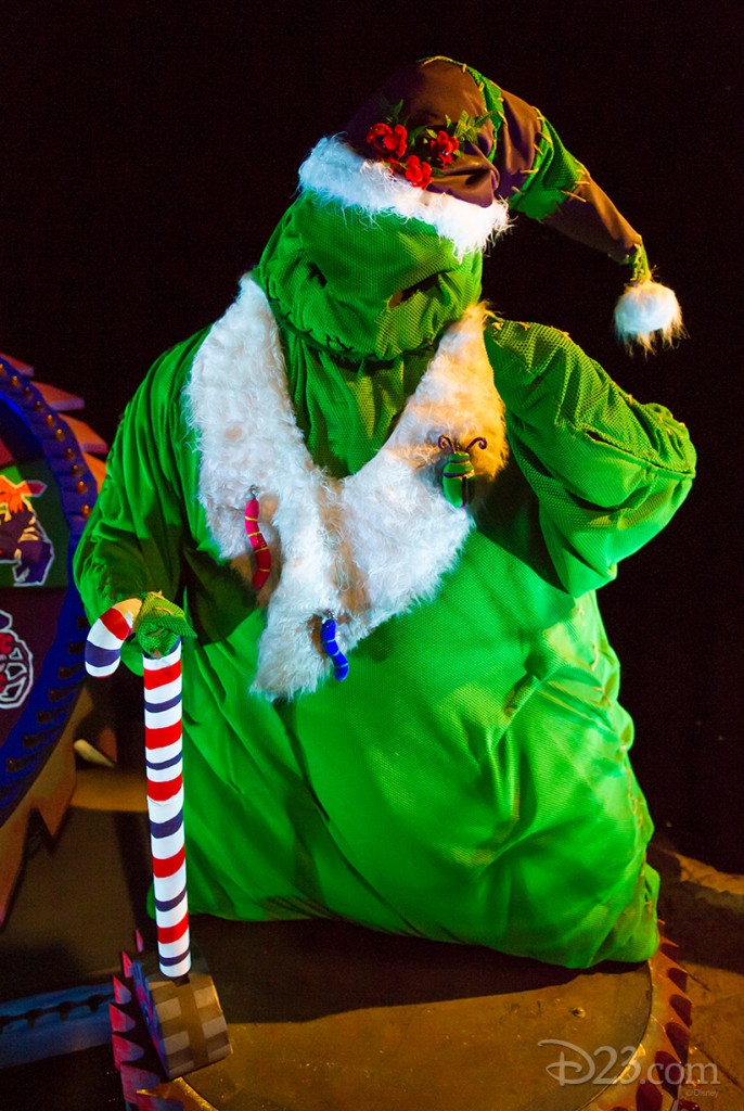 photo of character in bright green costume wearing white fur trimmed cap and coat and holding a large red-white-and-blue striped candy cane