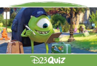 Mike Wazowski, a green, one-eyed monster from Monsters University, is looking at his check list in one hand and has a pen in his other hand while standing on a college campus walk path with green grass on either side of the path. He is wearing a dark blue hat that has "MU" written on the front of it while wearing a dark blue backpack with a tan suitcase and a green suitcase, both featuring stickers, on either side of him.