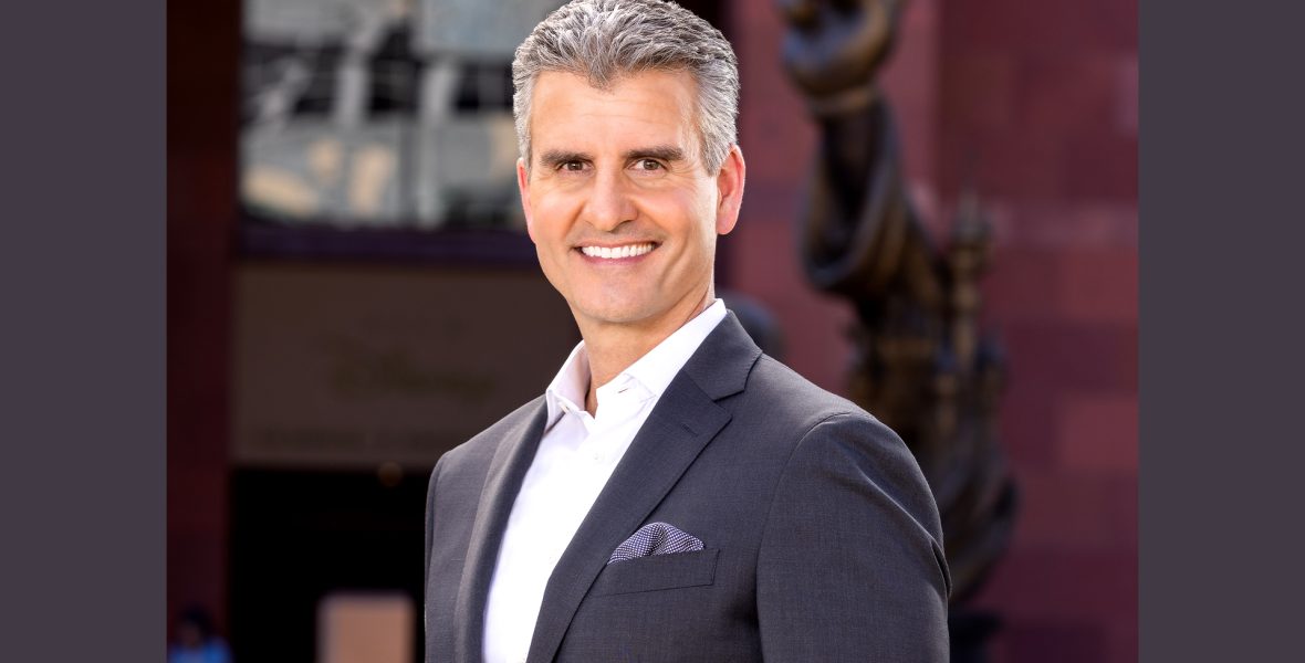 osh D'Amaro stands outdoors in a dark suit and white shirt with a Disney statue and building in the background.