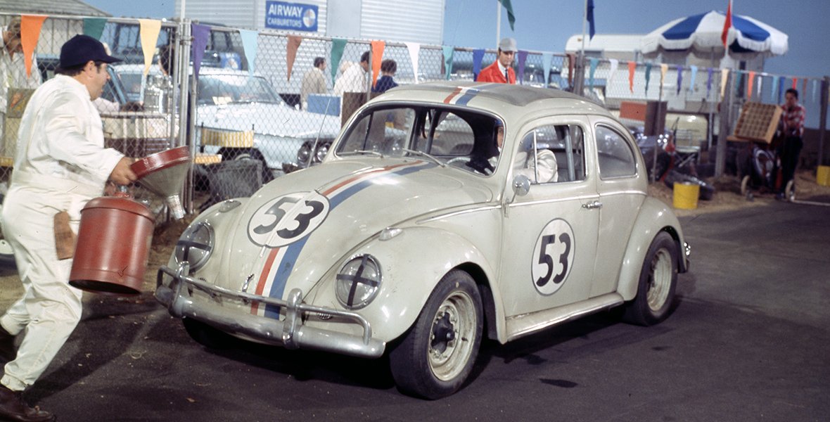 Herbie—a white love bug painted with red, white, and blue stripes and the number 53—is parked for a pit stop along a chain link fence. Tennessee, in a white jump suit, is running towards the car to service it before it resumes racing.