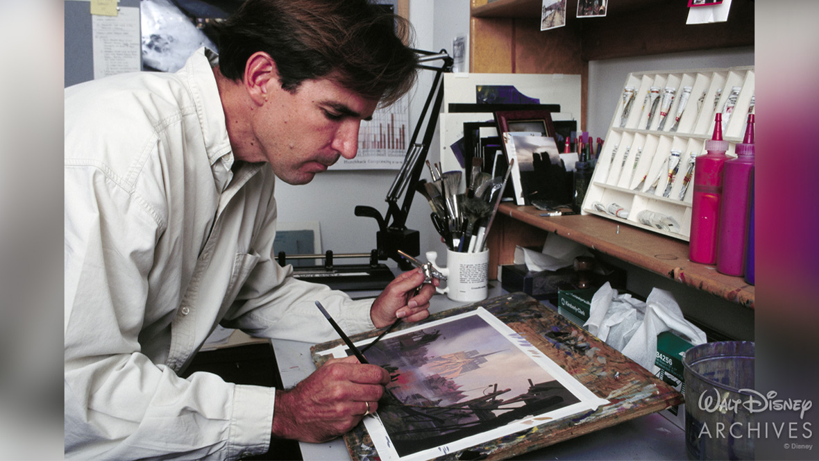 Art director David Goetz is stationed at a desk, painting a detailed scene on paper using a brush and an airbrush. The desk is surrounded by art supplies, brushes, and paint tubes.