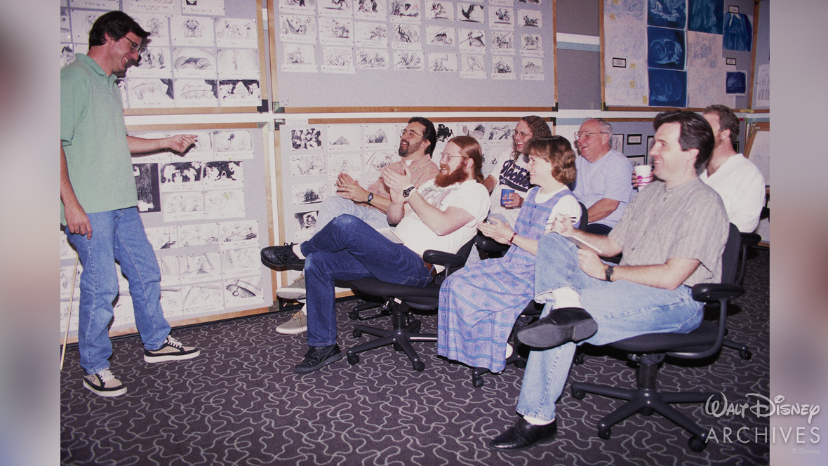 Members of the production team in casual clothing sit in rolling chairs, reviewing and discussing a wall full of storyboard sketches. Kevin Harkey stands and gestures toward the sketches while others look on and clap.
