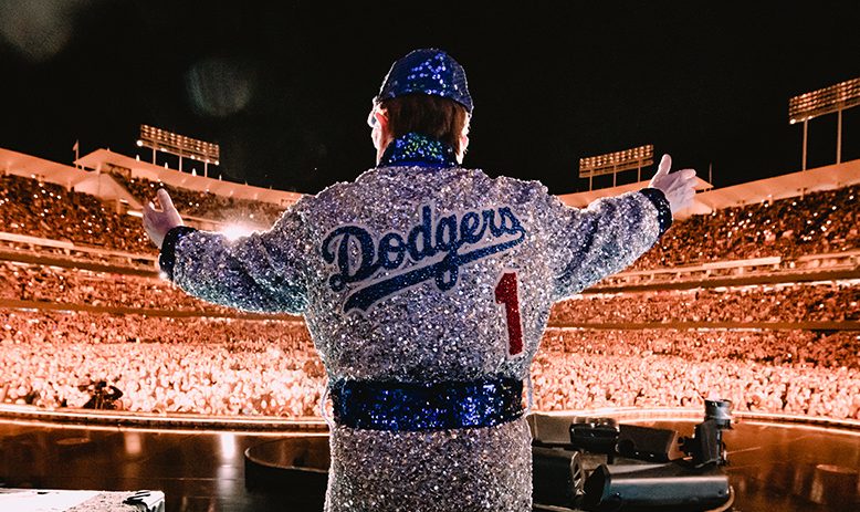 Elton John, wearing a bejeweled robe designed to mimic a Dodgers uniform, has his back to the camera, arms wide while facing a massive crowd inside Dodgers Stadium. 