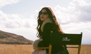 Olivia Rodrigo, in sunglasses and black coat with brown faux fur accents, sits in a director’s chair in a field. The back of the director’s chair is facing the camera and features the word “Sunset” in white text. Olivia has turned her body around in the chair so she is facing the audience. Behind her is a blue sky with fluffy white clouds.