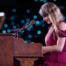 Taylor Swift, wearing a red dress, is playing a piano decorated with flowers. She is looking down at the keys with a smile on her face. Behind her, the concert crowd is out of focus, so that all we can see are blue dots of light in a bokeh effect against a black background.