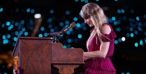 Taylor Swift, wearing a red dress, is playing a piano decorated with flowers. She is looking down at the keys with a smile on her face. Behind her, the concert crowd is out of focus, so that all we can see are blue dots of light in a bokeh effect against a black background.