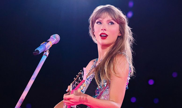 Taylor Swift, wearing a sparkly iridescent body suit, looks out into the distance while playing a pink guitar. A pink and purple microphone and stand is directed at her face, although she appears to not currently be singing. 
