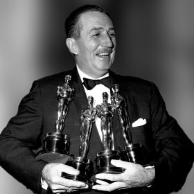 Walt Disney in a suit holds four oscars in his hands.