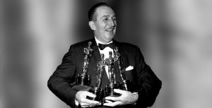 Walt Disney in a suit holds four oscars in his hands.