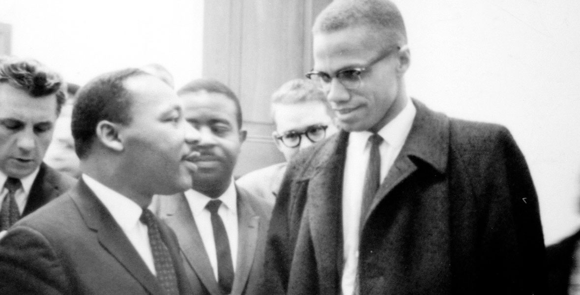 Black-and-white image of Dr. Martin Luther King Jr. and Malcolm X chatting, with several other men standing behind them.