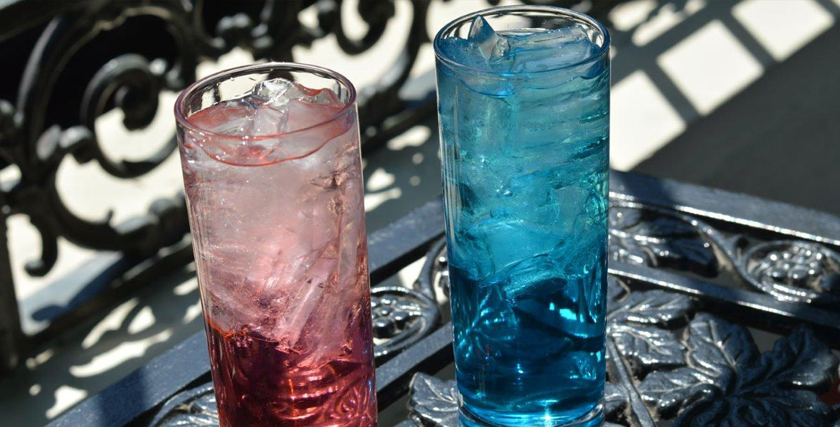 Side-by-side image of two Ralph Brennan’s Jazz Kitchen beverages—the red Rebel cocktail and the blue Empire cocktail; each is in a tall clear glass, with ice, set on a black wrought iron table. 