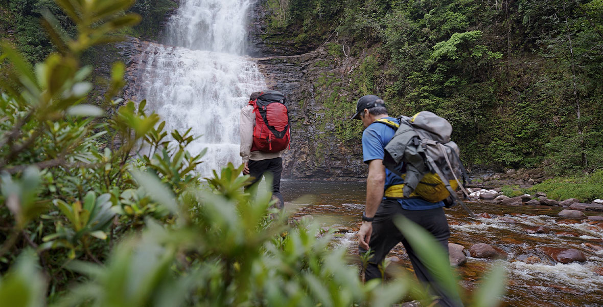 Two explorers walk through the river towards a waterfall surrounded by a lush forest.