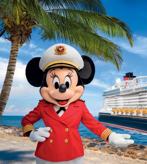 Captain Minnie stands in front of a palm tree with a Disney cruise ship behind her at sea.