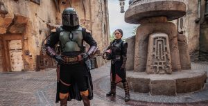 Boba Fett, with his helmet on, and Fennce Shand, holding her helmet in her arm, stand inside the Star Wars: Galaxy’s Edge marketplace.