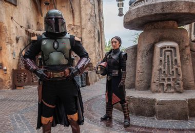 Boba Fett, with his helmet on, and Fennce Shand, holding her helmet in her arm, stand inside the Star Wars: Galaxy’s Edge marketplace.