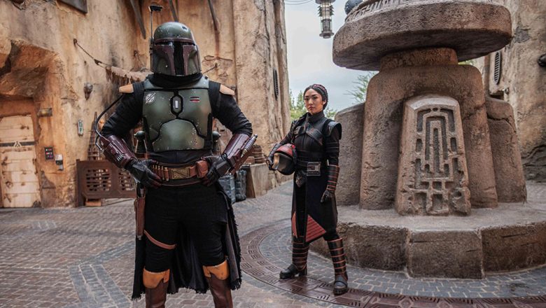 Boba Fett, with his helmet on, and Fennce Shand, holding her helmet in her arm, stand inside the Star Wars: Galaxy’s Edge marketplace.