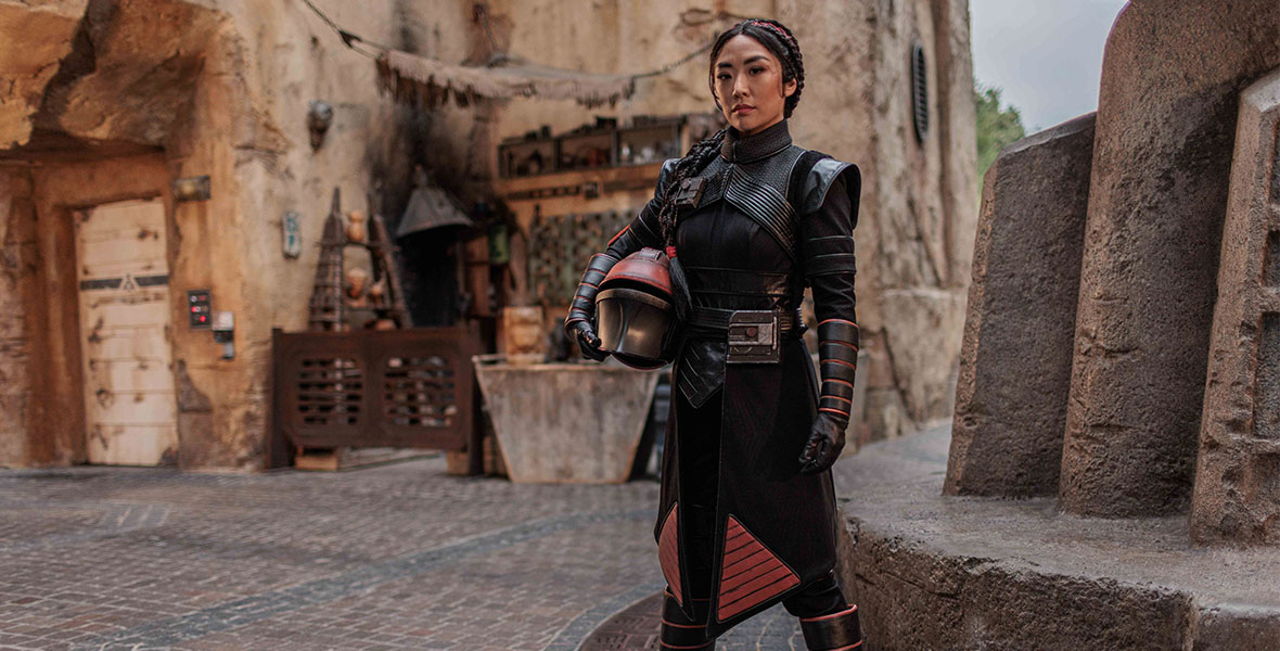 Fennec Shand, holding her helmet in her arms, stands inside the Star Wars: Galaxy’s Edge marketplace.