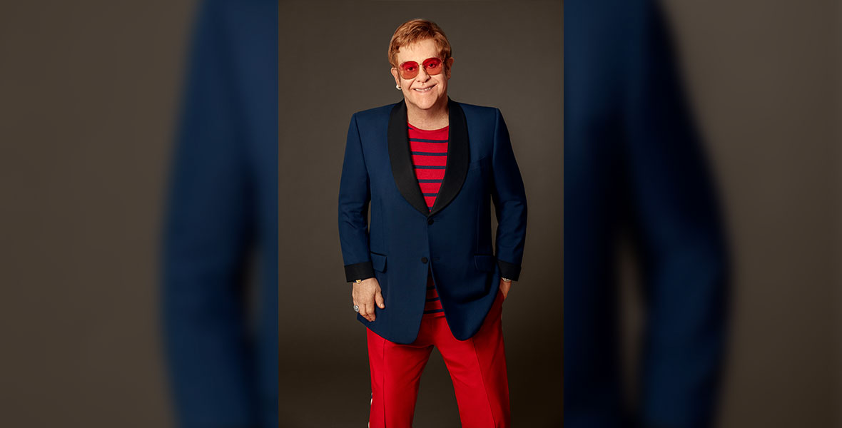 Disney Legend Elton John is standing in front of a brown background, wearing a dark blue blazer over a striped red-and-white shirt and red pants. He’s always wearing a signature pair of red eyeglasses.