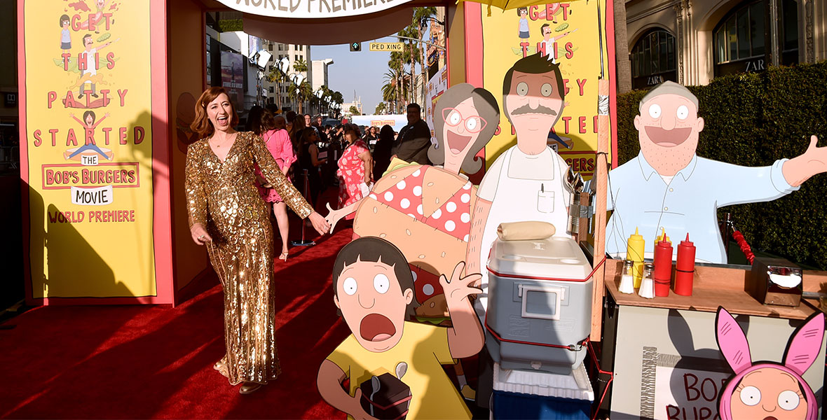 Kristen Schaal poses wildly wearing a gold, shiny ballgown near a cardboard cutout of the Belcher family on the red carpet at The Bob’s Burgers Movie world premiere.