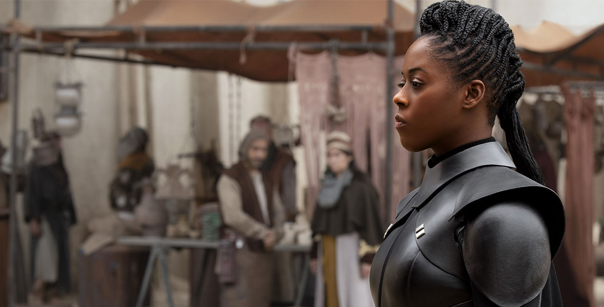 Moses Ingram, as Reva, walks through the streets of Tatooine in her black Inquisitor armor, a serious expression on her face. She is in profile, facing left.