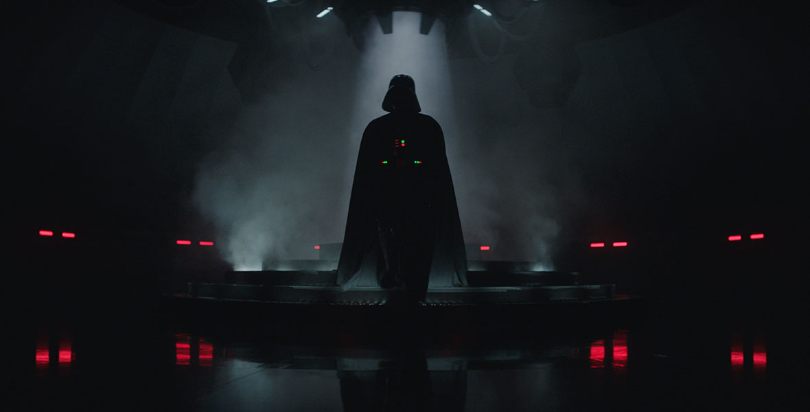 Darth Vader stands in silhouette in the center of the image. Everything around him is dark except for red lights along the walls, the lights on his armor, and the beam of light casting down on him from the ceiling.