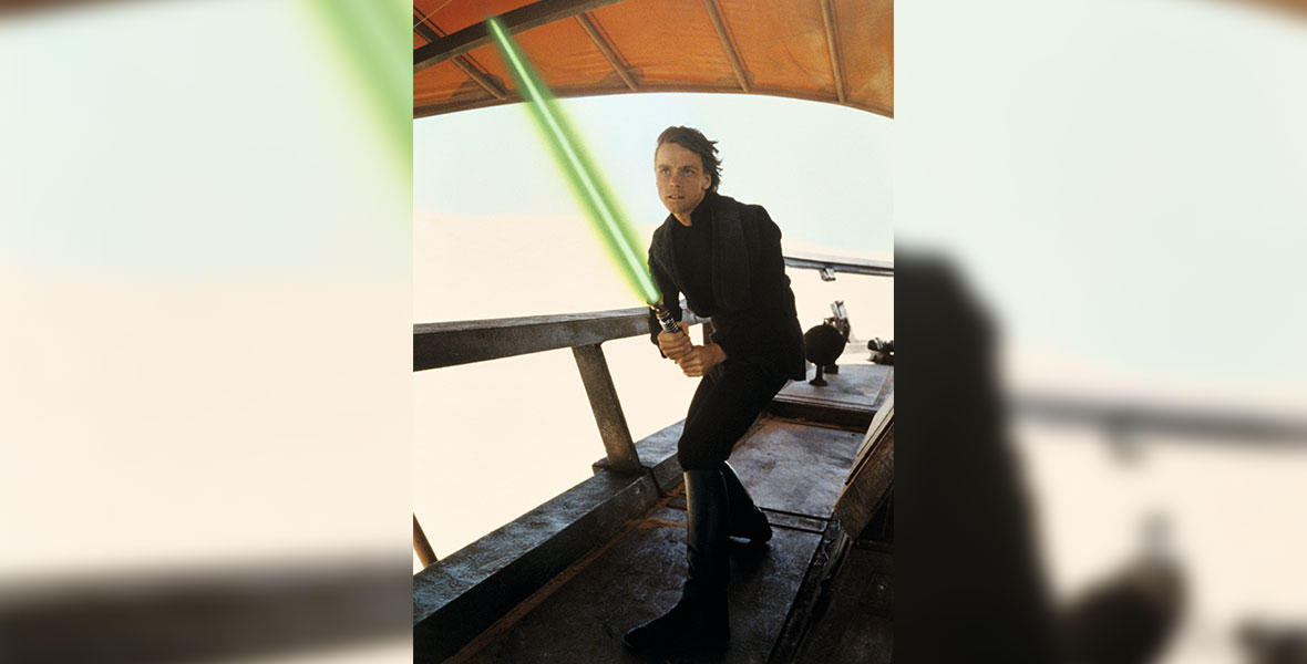 Luke Skywalker is posed to strike with his green lightsaber on one of Jabba the Hutt’s barges. He is wearing an all-black outfit and a focused expression.