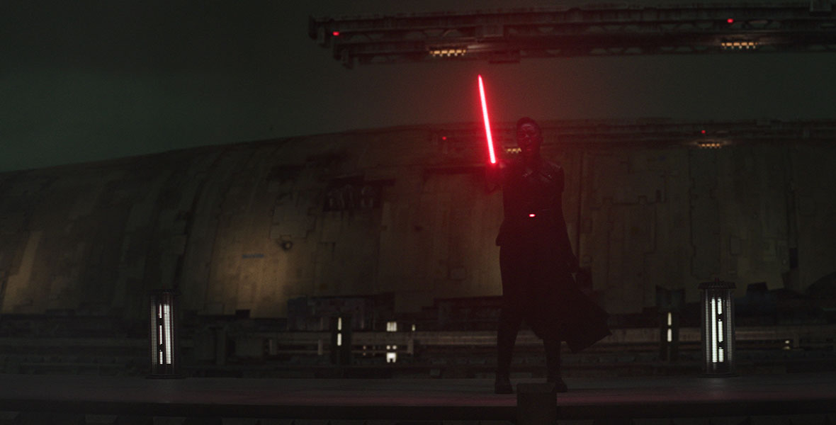 In darkness lit mostly by her red lightsaber, Reva brandishes her weapon and yells at someone or something offscreen.