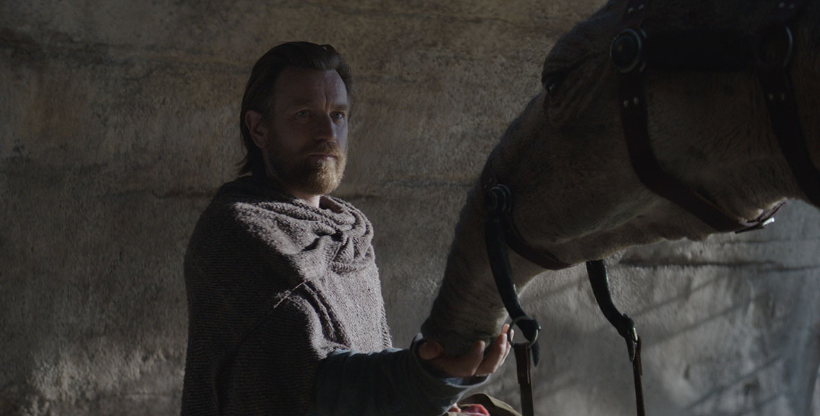Inside a cave, Obi-Wan holds a hand out to the snout of a large, alien creature wearing a bridle. His face is half-lit, and his expression is neutral.