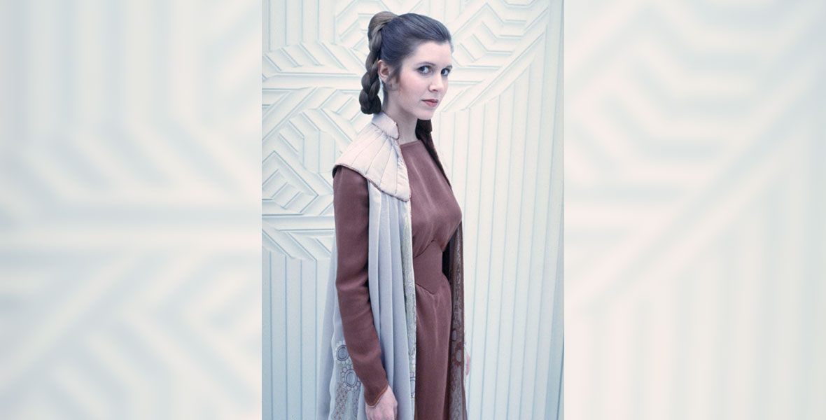 Princess Leia Organa looks into the camera with a neutral expression. She is against a white background and wearing a crimson dress and cape-like white vest.