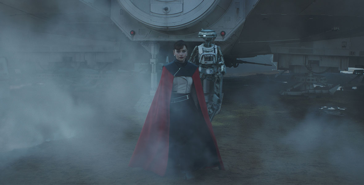 Qi’ra exits the Millenium Falcon with L3-37 behind her, both partially hidden by a thick fog. Qi’ra wears a dramatic red and black cape over practical clothes.