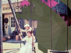 Sean Marshall (Pete) and Helen Reddy (Nora) sit on a swing in clean, fancy white outfits. They are being pushed on the swing by Elliot who looms enormously behind them.