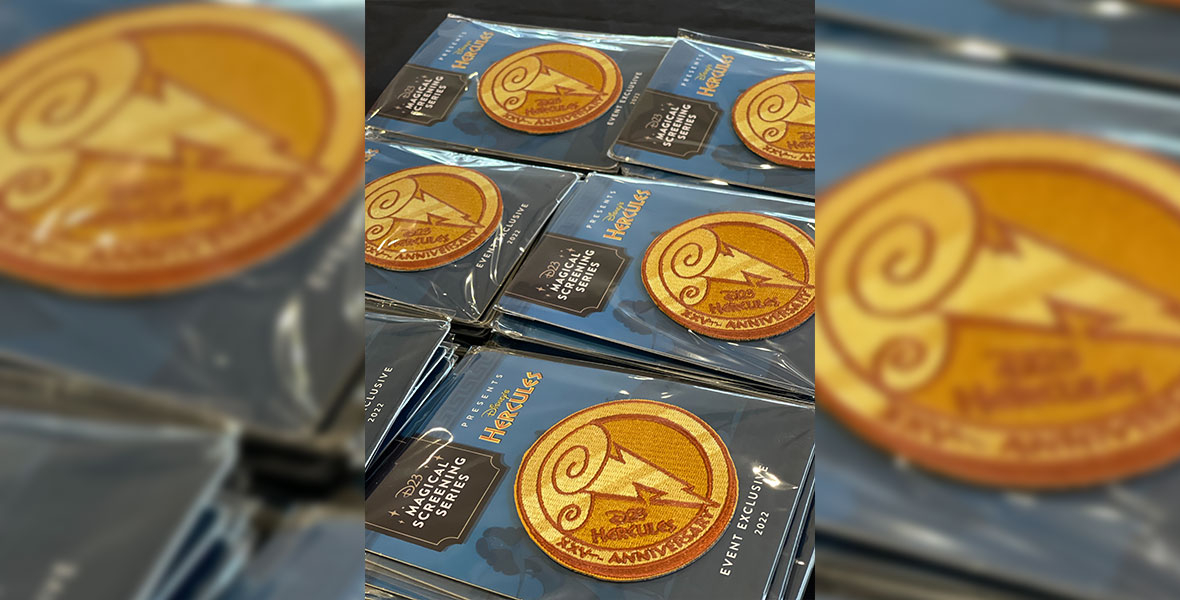 A close-up image of golden patches on a blue backing card. The round patch is styled as a medallion bearing a cloud and lightning bolt, along with the text “D23 Hercules— XXVth Anniversary”