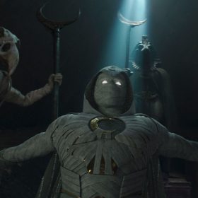 Marc Spector, in his full Moon Knight suit, kneels in a beam of light inside Khonshu’s tomb. Beside him, in partial darkness, is the god Khonshu himself.