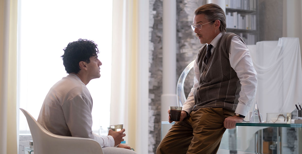 Marc Spector, dressed as a hospital patient, sits facing Arthur Harrow, dressed as a psychologist and leaning on a desk.