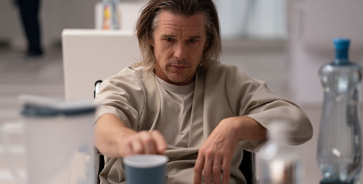 Arthur Harrow, dressed as a hospital patient, sits in a wheelchair and reaches for a blue cup that is slightly out of focus.