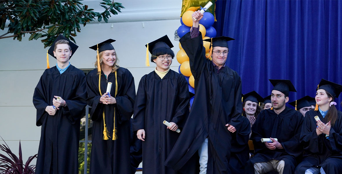 A colorful stage full of graduates both standing and sitting whilst wearing their cap and gown. One boy stands in the middle of stage holding his diploma in the air above his head.