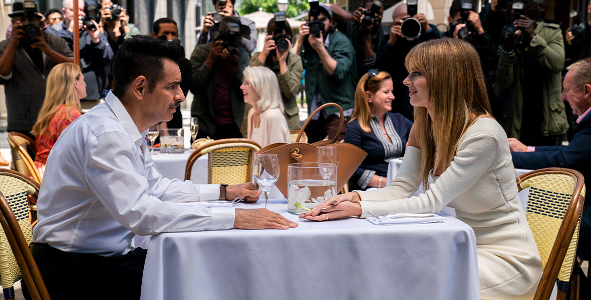 A man and a woman sit across from one another at an outdoor restaurant with paparazzi cameras surrounding the table.